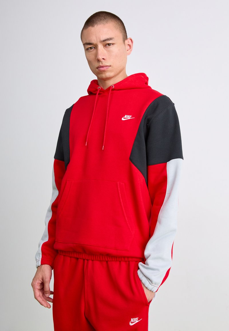 Tânăr îmbrăcat într-un hanorac Nike roșu, negru și alb, împreună cu pantaloni de trening Nike roșii asortați, stând în fața unui fundal deschis.