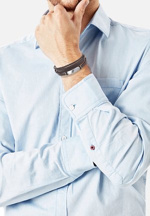 Bracciale in pelle e metallo con doppie cinghie in pelle nera e un accento metallico argentato, indossato su un polso accanto a una camicia azzurro chiaro.