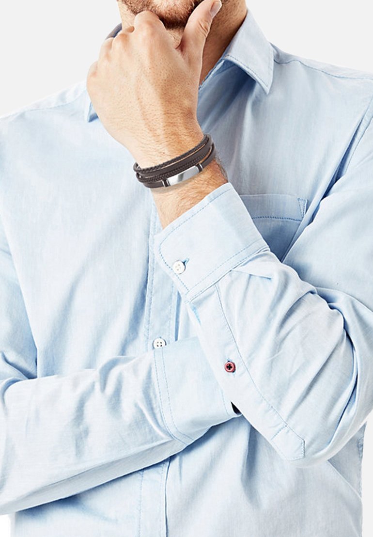 Pulsera de cuero y metal con doble correa de cuero negro y un acento metálico plateado, llevada en una muñeca junto a una camisa de color azul claro.