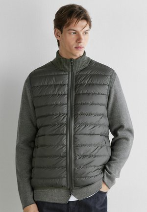 Junger Mann trägt eine dunkelolivgrüne gesteppte Weste mit Reißverschluss über einem grau gestrickten Pullover und schaut seitlich vor einer schlichten hellen Wand.