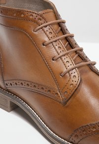 Chaussure habillée en cuir marron avec détails brogue, lacets ronds et talon en bois empilé sur fond blanc.