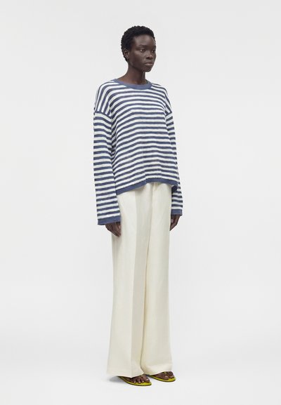 Γυναίκα στέκεται φορώντας μπλε και άσπρο ριγέ πουλόβερ, φαρδιά λευκά παντελόνια και κίτρινες σαγιονάρες σε μονόχρωμο φόντο.