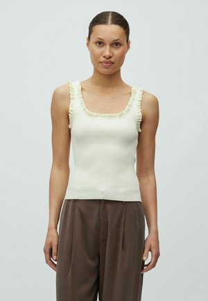 Femeie cu păr închis la culoare, purtând un top alb fără mâneci, din material nervurat, cu bretele volanate și pantaloni largi maro, în picioare pe un fundal simplu, deschis la culoare.