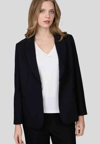 Blazer noir ajusté en tissu lisse, avec fermeture à un bouton, revers à crans et manches longues. Porté sur une chemise blanche à col en V.