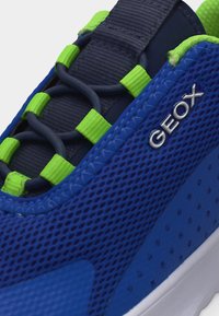 Κοντινό πλάνο σε μπλε αθλητικό παπούτσι με πλέγμα, φτιαγμένο με νεον πράσινους θηλές για κορδόνια, ναυτικομπλε ελαστικά κορδόνια και ασημένιο λογότυπο "GEOX" στο πλάι.