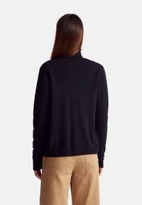 Černý svetr s vysokým límcem, dlouhými rukávy a žebrovaným lemem, kombinovaný s béžovými kalhotami s zadními kapsami a volným střihem.