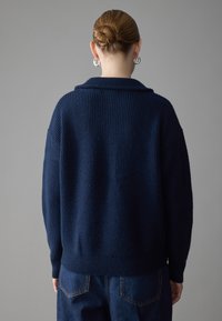 Cardigan in maglina blu navy con collo sciallato, taglio oversize, texture a costine e spalle scese, abbinato a jeans in denim scuro.