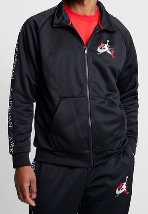 Mężczyzna w czarnej rozpinanej kurtce Nike Air Jordan i pasujących spodniach, pod spodem czerwona koszulka, loga na piersi, rękawie i spodniach.