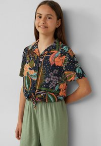 Kurzärmliges Hemd mit einem dunklen Blumenmuster, das leuchtend orangefarbene und grüne Akzente aufweist. Kombiniert mit hellgrünen, locker sitzenden Hosen.