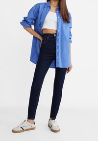 Blue button-up shirt with rolled sleeves over a white cropped tank top, paired with dark high-waisted jeans and beige sneakers.