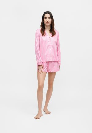Femme debout pieds nus portant une chemise rose rayée à manches longues boutonnée et un short assorti à coupe décontractée sur fond uni.