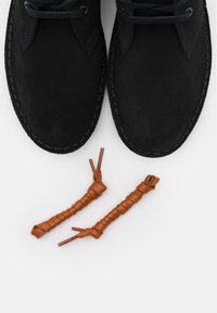 Schwarze Wildlederstiefel bis zum Knöchel mit runden Schnürsenkeln. Die Schnürsenkel werden durch zwei braune Seilschnürsenkel mit gewickelter Textur ergänzt.