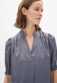 Harmaa satiini-blussi, jossa on lyhyet puff-hihat, napittomalla V-kaula-aukolla ja kootulla yksityiskohdalla hihojen helmassa. Tasainen rakenne, jossa on hillitty kiilto.