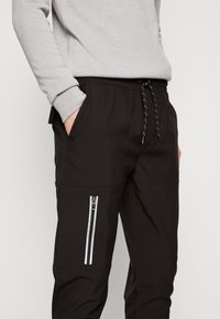 Person som bär svarta dragsko-byxor med sidoficka med dragkedja och händerna i framfickorna, tillsammans med en ljusgrå långärmad topp.