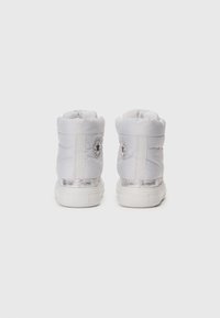Botines tobilleros blancos, acolchados y con una textura suave, que presentan un logotipo circular a cada lado y un acento metálico plateado alrededor de la suela.