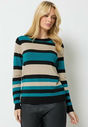 Donna con lunghi capelli biondi che indossa un maglione a righe verde acqua, beige e nero a maniche lunghe e pantaloni neri, in piedi davanti a uno sfondo semplice.