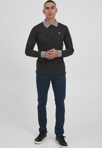 Langermet svart polo skjorte med grå krage og mansjetter, med et lite emblem på brystet, kombinert med marineblå bukser og svarte joggesko.