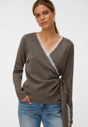Femme portant un pull cache-cœur marron avec une bordure en dentelle blanche, associée à un jean bleu, posant devant un fond uni.