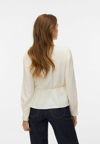 Bluzka w kolorze kremowym z długim rękawem o fakturowanej tkaninie, z marszczonym pasem i delikatnie bufkami na rękawach, zestawiona z ciemnymi dżinsami.