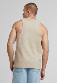 Beige geribbelde tanktop met een ronde halslijn, gekenmerkt door een textuurstof. De achterkant heeft een subtiele merklabel en een relaxte pasvorm.