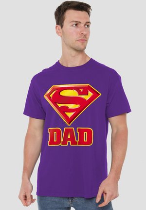 Man wearing purple T-shirt with red and yellow Superman-style emblem above the word "DAD," standing against a plain background.