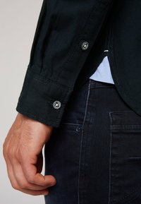 Puño de camisa negra texturizada con detalle de botón, combinado con jeans de mezclilla oscuros. La camisa presenta costuras en contraste y un aspecto en capas.
