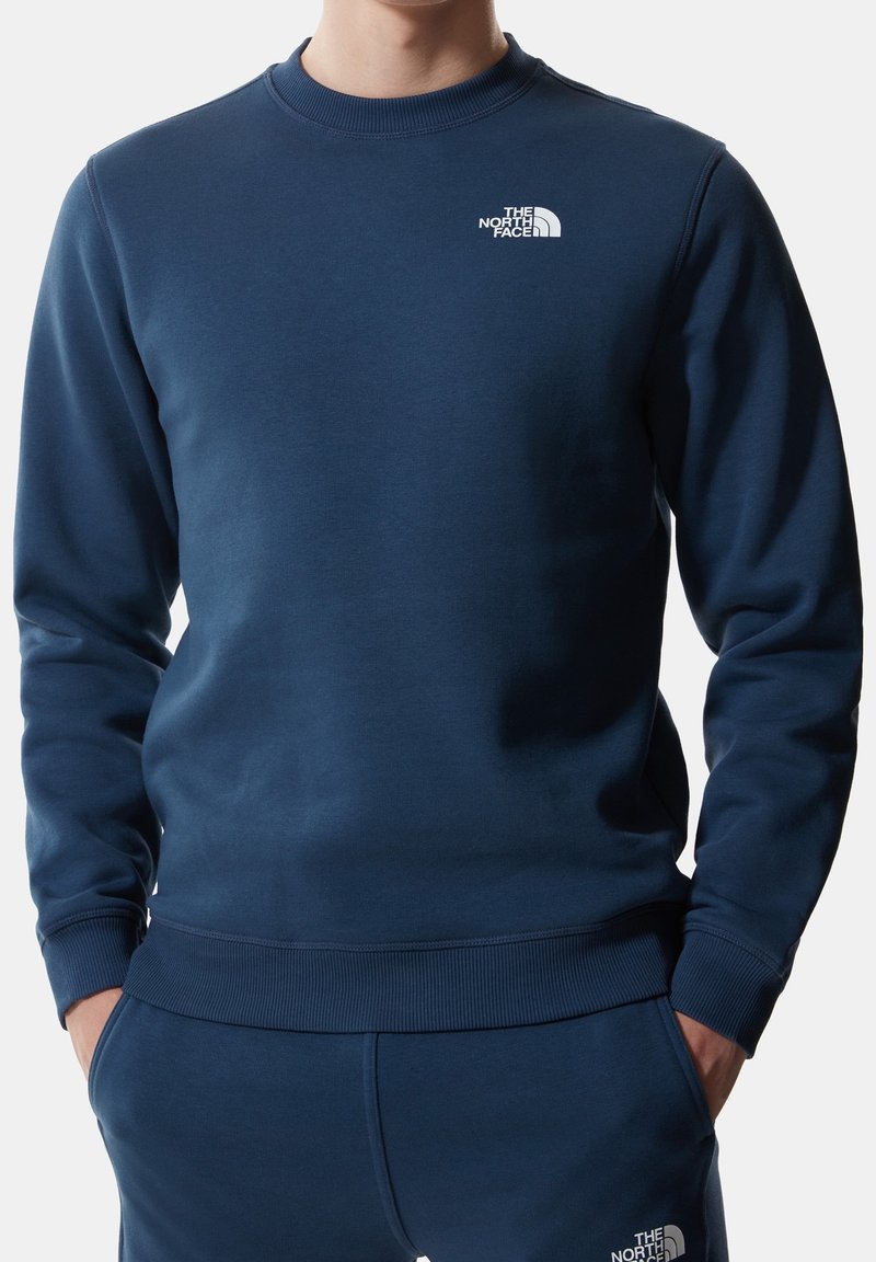 Homme portant un sweat-shirt bleu marine et un pantalon assorti avec le logo The North Face sur la poitrine et la cuisse, les mains dans les poches.