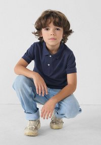 Marineblauwe poloshirt met korte mouwen, twee knopen en geborduurd logo, gecombineerd met lichtblauwe jeans en beige sneakers.
