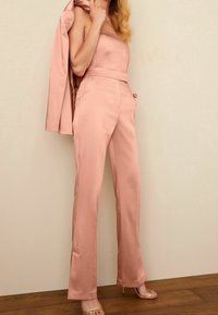 Różowy satynowy kombinezon z tapered pants i dopasowanym górnym elementem, zestawiony z pasującą marynarką zarzuconą na jedno ramię. Złote sandały na obcasie.