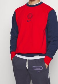Sweat-shirt rouge et marine à encolure ronde, avec un graphique de basketball et le logo Nike. Pantalon de survêtement gris avec détail "LIVERPOOL FC" en violet.