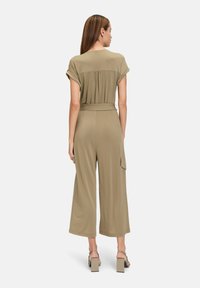 Olivgrön jumpsuit med vikta ärmar, åtdragen midja och bredbyxdesign. Har en bakfåll och sidofickor för cargo. Slät textur.