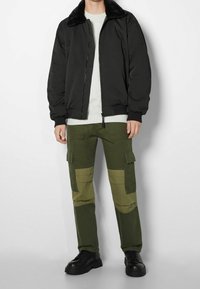 Svart bomberjacka med faux fur-krage, ljusgrå sweatshirt och gröna cargobyxor med överläggsfolie. Svarta sneakers fulländar outfiten.