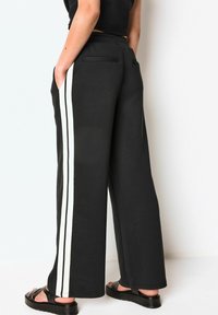 Pantaloni neri a gamba larga realizzati in tessuto liscio, con due strisce bianche sui lati e tasche laterali con tasche posteriori a toppa, abbinati a sandali neri.