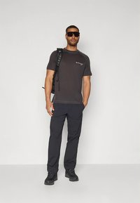 Dunkelgraues T-Shirt mit kurzen Ärmeln, schwarze Cargo-Hose, schwarze Wanderschuhe und ein Rucksack. Zubehör umfasst eine Sonnenbrille und ein minimalistisches Logo.