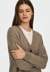 Brauner gestrickter Cardigan mit V-Ausschnitt, schwarzer Knopfleiste und gerippten Bündchen. Getragen über einem weißen T-Shirt. Glatte Textur.