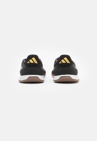 Zapatillas negras con parte superior de tela texturizada, detalles dorados en el talón y una entresuela blanca con una suela de goma marrón.