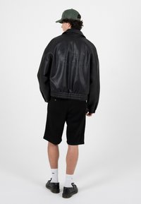 Muž v černé kožené bundě, černých šortkách, bílých ponožkách, černých botách a zelené baseballové čepici, otočený zády proti jednobarevnému bílému pozadí.