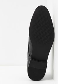 Semelle de chaussure noire en caoutchouc texturé, bord arrondi et logo gravé. Dessus lisse avec une légère courbe au talon, design minimaliste.