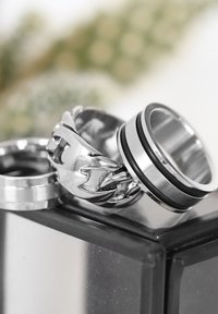 Three metallic rings are displayed: a polished band, a textured chain design, and a two-tone ring with a black stripe.