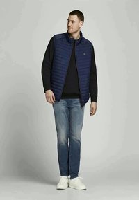 Gilet trapuntato blu navy sopra un maglione nero, abbinato a jeans azzurri chiari e sneakers bianche. Presenta colletto alto e chiusura con zip.