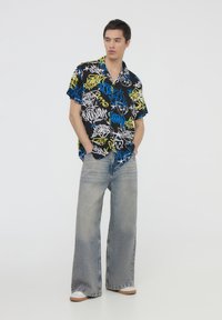 Maglietta nera a maniche corte con motivi graffiti colorati, abbinata a jeans larghi blu chiaro e sneakers bianche con suola beige.