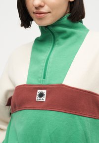 Femme portant un pull vert à col montant, avec une bande bordeaux sur la poitrine, des manches crème et un patch logo soleil noir sur la poitrine.