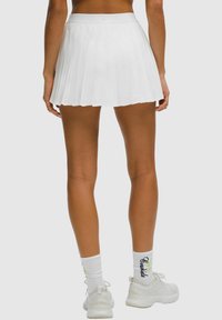Woman wearing a white pleated mini skirt, white branded socks, and white sneakers standing against a neutral background.