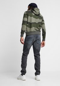 Homme portant un sweat à capuche camouflage vert, un bonnet noir, un jean gris foncé et des baskets blanches, debout dos à nous sur un fond uni.