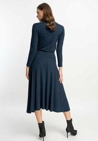 Marineblaue Langarmkleid mit tailliertem Oberteil und schwingendem Rock, aus weichem Stoff. Kombiniert mit schwarzen Knöchelstiefeln. Einfaches, modernes Design.