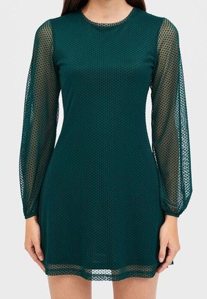 Robe vert foncé à manches longues transparentes avec une texture à petits points, portée par une personne debout avec les mains détendues le long du corps.