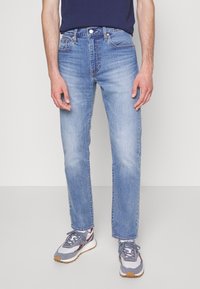 Pantalones vaqueros de mezclilla azul claro con corte recto, cinco bolsillos y remaches de cobre. Combinados con zapatillas grises con detalles en blanco y rojo.