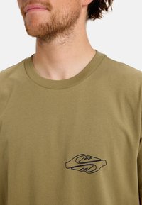 Camiseta de algodón color verde oliva con cuello redondo. Presenta un diseño gráfico azul en la parte izquierda del pecho y una textura lisa y sencilla.