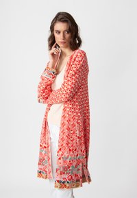 Garšs kardigans korallkrāsā ar ģeometriskiem rakstiem. Iezīmējas ar vieglu audumu, atvērto fronti un krāsainiem akcentiem piedurkņu un apakšmalas daļā.