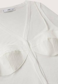 Top a V con scollo a V in tessuto bianco a costine, caratterizzato da due accenti curvi sovrapposti sul petto. Texture liscia con design aderente. Etichetta del brand visibile.